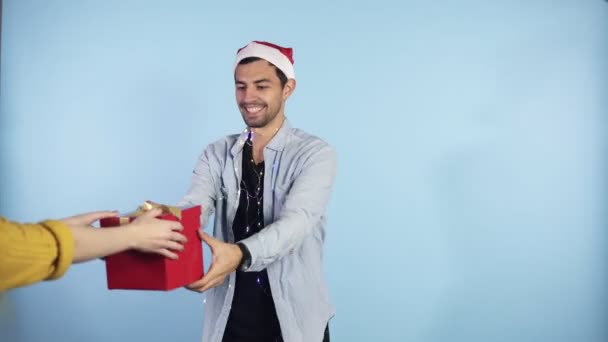 Pozitivní, mladý muž ve vánočním klobouku a věnci na krku dostává překvapení z boku ženské ruce - je v dobré sváteční náladě, izolované přes modré pozadí. Lidé, překvapení a dárky — Stock video
