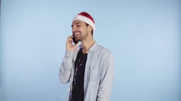 Giovane uomo emotivo felice indossa Natale cappello di Babbo Natale e ghirlanda sul collo in piedi e parlando con il telefono cellulare. Uomo in casuale isolato su sfondo blu parete — Video Stock