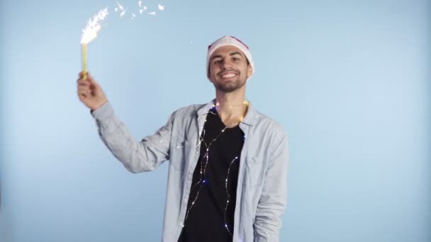 Un bel homme avec un chapeau rouge de Noël et une guirlande sur le cou danse et agite la main avec un craquelin brûlant, célébrant Noël ou le Nouvel An. Fond isolé sur bleu — Video