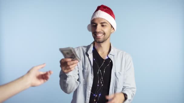 Joven hombre feliz recibió papel moneda, billetes de dólar. Contando el dinero mientras está de pie contra la pared azul en sombrero de santa y guirnalda en el cuello. Obtener dinero en efectivo, recibir el pago del ganador, concepto de regalo. Hombre. — Vídeo de stock