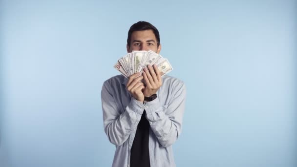 Astonished caucasian man in casual clothes holding bunches of dollars, lucky lottery winner, super excited about it. Holding paper money before face, cant believe in this luck. Blue wall background — ストック動画