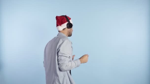 Pozitív jóképű férfi egy karácsonyi piros sapkában fejhallgatóval, kedvenc zenéjét hallgatva és táncok egy gondtalan ritmusban - kék fal háttér stúdió. Férfi alkalmi ruhák és kalap szabadon — Stock videók