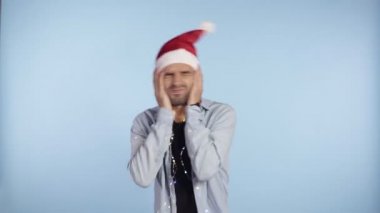 Geleneksel Noel Baba şapkalı genç çılgın adam başını sallıyor, deliye dönüyor. El ele tutuşuyor, yüzünü buruşturuyor ve mavi arka planda tek başına duruyor.