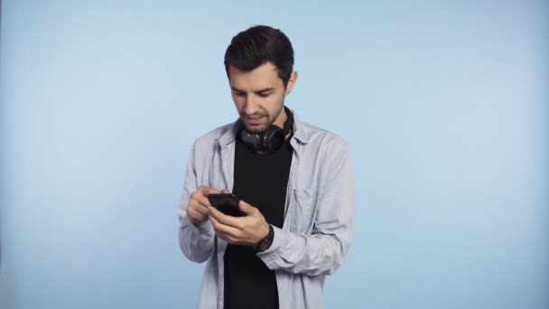 Красивый, любопытный мужчина в голубой футболке и наушниках на шее представляет смартфон и показывает на камеру смартфон с зеленым экраном, изолированный на синем фоне — стоковое видео