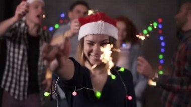 Boyunda renkli ışıklar ve kameraya poz veren Noel Baba şapkalı güzel bir kız. Arkadaşları dekore edilmiş odada bulanık arka planda dans ederken gülümsüyor, bengal ışığını sallıyor. Arkadaşlar kutluyor