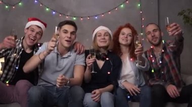 Happy young people drinking champagne from the glasses during celebrate New Year or Christmas Eve, having great time in relaxing home atmosphere with garlands lights everywhere. Group of young friends
