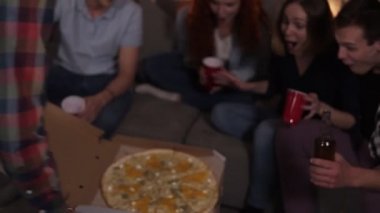 Happy friends got delivery pizza and celebrating party clinking bottles and red cups with beer and soda sitting on sofa at home. So excited about big pizza party