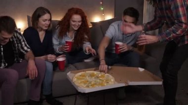 Diverse people, friends got together to eat pizza at home. Got delivery Italian pizza with cheese, taking slices, holding red cups and bottles. Starting home pizza party. Garlands light on the