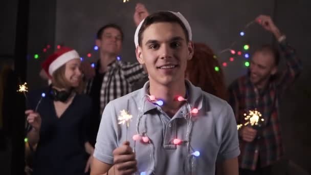 Portrait of caucasian young guy with colorful lights on neck and santa hat posing for camera - smiling, holding his bengal light while his friends dancing and celebrating on the blurred background in — Wideo stockowe