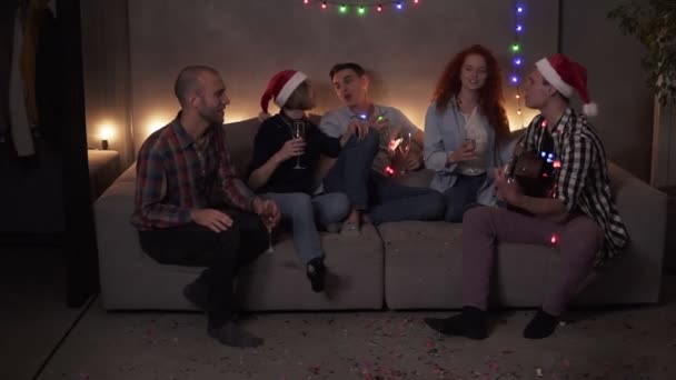 Alegre amigos cercanos celebrando, tener tiempo juntos en casa. Amigos felices se sientan en el sofá y escuchan a un tipo cantando y tocando la guitarra. Reúnanse en Navidad. Luz apagada, guirnalda — Vídeo de stock