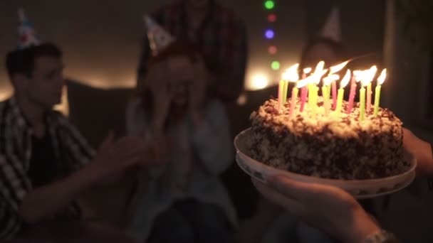 Szczęśliwa uśmiechnięta rudowłosa dziewczyna zdmuchuje świeczki na swoim małym torcie urodzinowym. Dziewczyna otoczona przez najbliższych przyjaciół. Światła przytłumione, światła girlandowe — Wideo stockowe