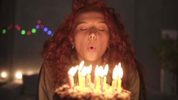 Retrato de uma menina encaracolada encaracolada sorrindo fazendo um desejo e soprando velas em seu bolo de aniversário. Luz abafada, luzes de guirlanda. Movimento lento — Vídeo de Stock