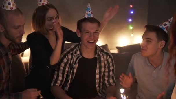 Renkli şapkalı arkadaşlar doğum günü partisini kutluyor, sürpriz bir arkadaş elinde yanan sopayla pasta yapıyor. Çok heyecanlı bir adam gülüyor. Tavandan parlayan konfeti düşüyor. En yakın arkadaşlar — Stok video
