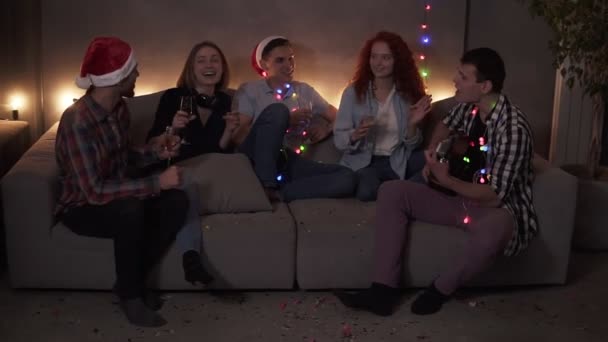 Cheerful close friends celebrating, having time together at home. Happy friends sit around on sofa and listen to guy singing and playing guitar. Get together at Christmas time. Muffled light, garland — Stock videók