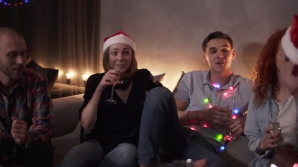 Feliz Año Nuevo Grupo de personas celebrando, tener tiempo juntos en casa. Amigos felices se sientan en el sofá bebiendo y cantando juntos baladas, hombre tocando la guitarra. Luz apagada, luces de guirnalda — Vídeo de stock