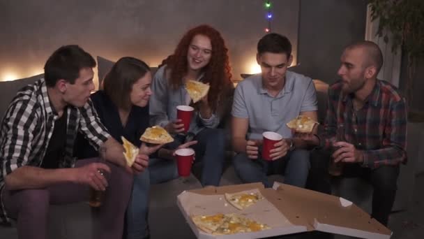 A fiatalok vidám férfiak és nők giccses pizzát esznek, beszélgetnek pihentető közben a lakásban, egy tetőtéri szobában. Ízletes pizza kartondobozban látható az asztalon, kezében palackok és poharak — Stock videók