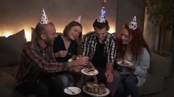 Θετικοί τύποι στο σκοτάδι, διακοσμημένοι με γιρλάντες, κάθονται στον καναπέ και τρώνε εορταστική τούρτα γενεθλίων. Ο εορτάζων βάζει κομμάτια νόστιμης τούρτας σε πιάτα φίλων. Γιορτή γενεθλίων με — Αρχείο Βίντεο