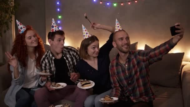 Wesoły, młody przyjaciel lub dwie pary mające małe, przytulne urodziny, siedzące razem w świątecznych stożkach na kanapie i robiące selfie przy użyciu telefonu komórkowego. Płyty z tortem urodzinowym — Wideo stockowe