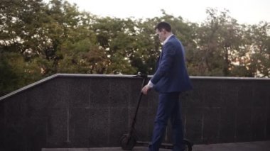 Elektronik scooter kullanan bir iş adamı. Telefonlara bakmak için duruyor. Güler yüzlü işadamı sokakta scooter sürüyor ve güneşli bir günde akıllı telefon kullanıyor. Ulaşım, hareketlilik kavramı