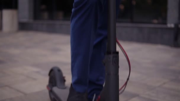 Imágenes hacia arriba de un joven hombre de negocios con elegante traje azul parado al aire libre, sonriendo alegremente y mostrando el pulgar hacia arriba. Hombre de negocios guapo con su scooter eléctrico en frente del edificio de negocios — Vídeos de Stock