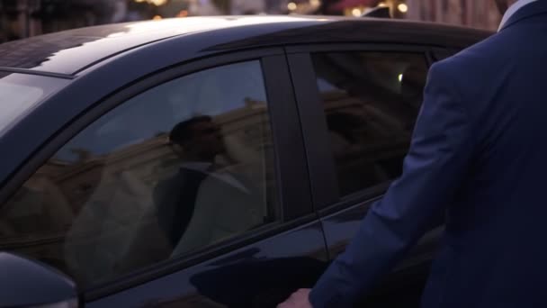 Slow motion jongeman lopen zittend in de zwarte auto op straat. Glimlachende zakenman in elegant blauw pak opent de deur van zijn dure auto en zit erin. Opgewonden sluit de deur. Avondschemering — Stockvideo