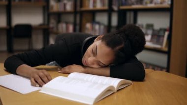 Siyah kazak giyen Afro-Amerikan bir bayan öğrenci, yorgun ve modern kütüphanede kitap ve notlarla dolu bir masada uyuyakalmış. Eğitim, insanlar, öğrenci ve öğrenme kavramı. Ön görünüm