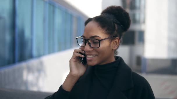 Mujer de negocios alegre en gafas hablando por teléfono inteligente. Emocional joven mujer de negocios afroamericana hablando por teléfono celular estar fuera en la calle. Concepto de conexión — Vídeo de stock