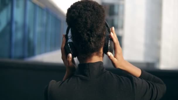 Seltener Blick auf junge attraktive schwarze Mädchen im Hintergrund moderner Stadtgebäude, die mit großen schwarzen Kopfhörern Musik hören. Frau in schwarzer Freizeitkleidung und Sonnenbrille. sonniger Tag — Stockvideo