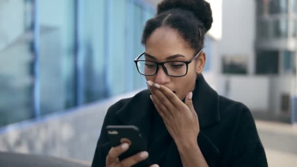 Shocked mujer afroamericana gots mensaje de ex, jadeos de sorpresa y emoción, lee noticias impresionantes en el teléfono móvil. Chica emocionada de pie al aire libre, en la calle. Vestido con ropa negra y — Vídeos de Stock