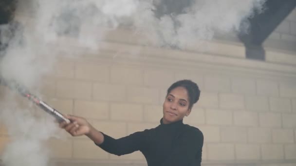 Jovem mulher afro-americana bonita duas bombas de fumaça ou granadas cores preto e branco no fundo da parede de tijolos brancos. Menina de camisola preta fazendo movimentos redondos com uma mão e dançando relaxantemente — Vídeo de Stock