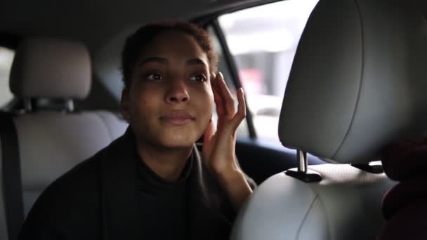 Jonge mooie Afro-Amerikaanse vrouw kijkt in achteruitkijkspiegel terwijl ze op de achterbank zit. zelfverzekerde vrouw in zwart kleding kijkt in de achteruitkijkspiegel slimmer omhoog — Stockvideo