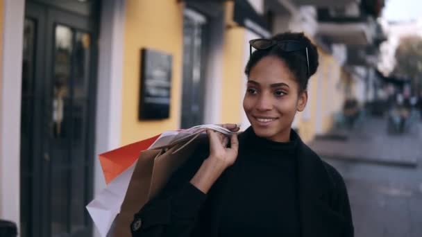 Portrait of an attractive mixed race girl smiling while walking down the street with colorful shopping bags. Happy young woman walking after shopping by fancy streets city, window shopping — ストック動画