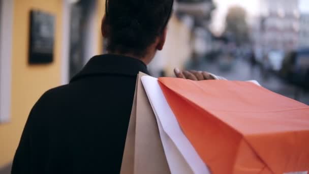 Vista rara de uma menina afro-americana andando pela rua com sacos de compras coloridos em um ombro. Despreocupado jovem mulher andando depois de fazer compras por ruas extravagantes, fundo borrado — Vídeo de Stock