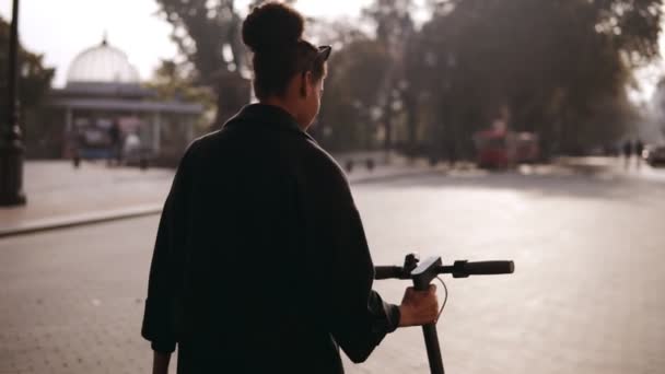 Mélange a couru femme marchant le long de la rue de la ville. Fille en manteau noir marchant avec scooter électrique coup de pied par la ville du matin. Au ralenti. Images de vue rares — Video