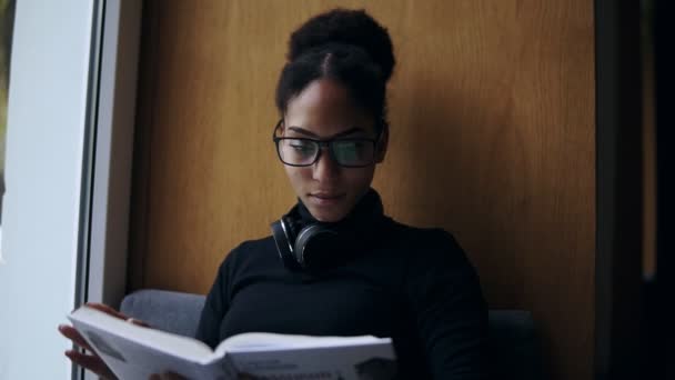 La joven y atractiva mujer afroamericana está leyendo el libro en el gran alféizar de la ventana. Retrato de cerca de una chica con estilo en gafas y auriculares leyendo o estudiando. De cerca. — Vídeo de stock