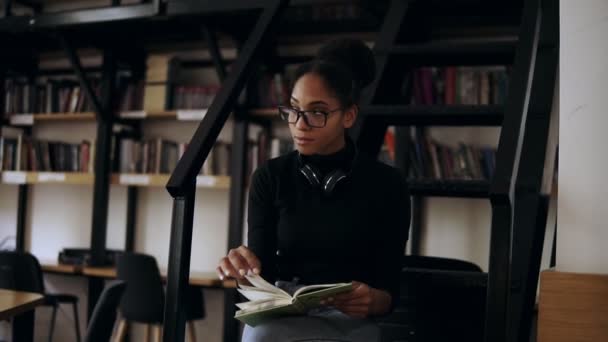 在现代图书馆，聪明的非洲裔美国学生从不同的书籍中收集信息，穿着黑色毛衣、头戴耳机的时髦女孩绕着脖子为书籍收集数据 — 图库视频影像