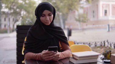 Açık kafedeki rahatlatıcı atmosferde siyah tesettürlü genç Müslüman kadın. Oturup ekranına bakıyor, sohbet ediyor, mesajları okuyor. Güzel, sıcak bir gün.