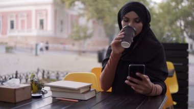Güzel, genç Müslüman bir kadın hafta sonları bir kafede akıllı telefondan sohbet ediyor ve kahve içiyor. Tesettürlü gülümseyen Müslüman kadın cep telefonu ve kahve fincanıyla mutlu bir şekilde oturuyor.