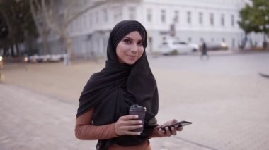 Mükemmel makyajlı, tesettürlü, kahve içen ve cep telefonu ekranına bakıp kameraya gülümseyen çekici genç bir kadın. Gerçek zamanlı görüntüler takip ediliyor