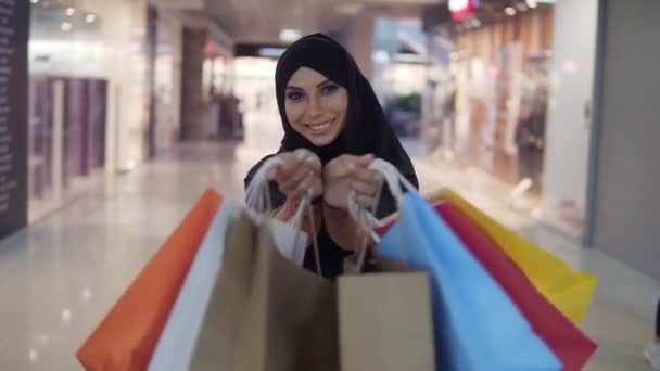 Mujer musulmana bastante joven con maquillaje perfecto sosteniendo varias bolsas de compras de colores después de ir de compras y sonriendo a la cámara. Vista frontal — Vídeos de Stock