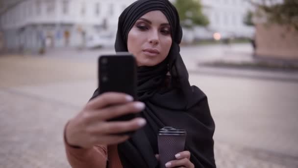 Mujer joven en hiyab negro con un maquillaje perfecto caminando, bebiendo café. Mujer bonita de pie fuera en la calle y haciendo selfie usando su teléfono inteligente — Vídeo de stock