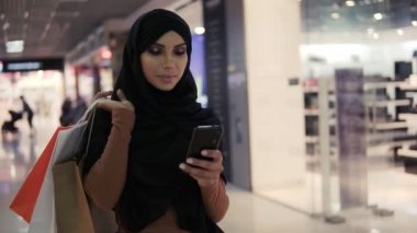 Siyah başörtüsü takan çekici Müslüman kadın alışveriş merkezinde yürüyor ve akıllı telefon kullanıyor. İletişim, çevrimiçi alışveriş, sosyal ağ kavramı