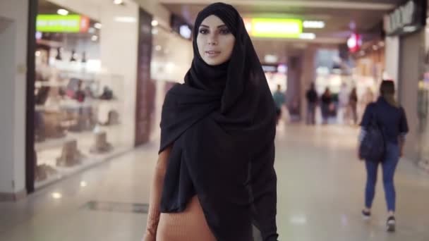 Portret van een stijlvolle jonge moslim vrouw in zwarte hijab en strakke bruine jurk met tassen in haar handen na het winkelen lopen door winkelcentrum. Langzame beweging. Arabische vrouw genieten van winkeltijd, glimlachen — Stockvideo