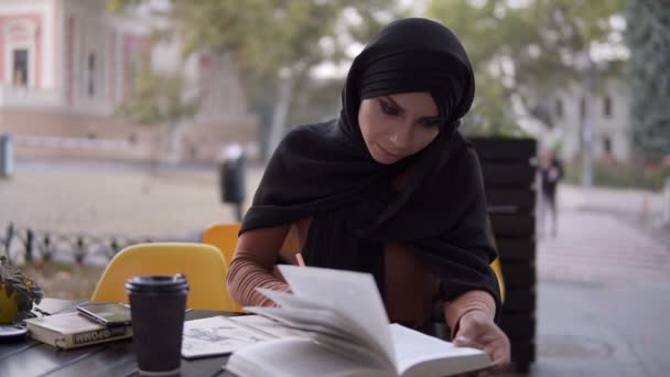 Una chica musulmana atractiva escribe en un cuaderno sentado en un café con un libro. Estudiando en la cafetería al aire libre, tomando un descanso. Universidad, autoeducación. Vista frontal — Vídeo de stock