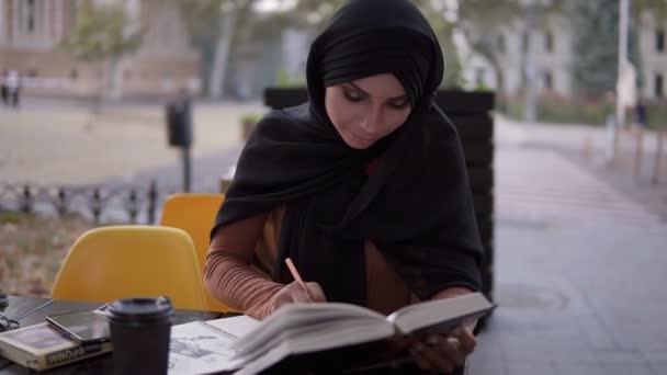 Seorang gadis muslim yang menarik menulis dalam sebuah notebook duduk di sebuah kafe dengan sebuah buku. Belajar di luar ruangan kafe, istirahat. Kuliah, pendidikan sendiri. Pemandangan depan. Gerakan lambat — Stok Video