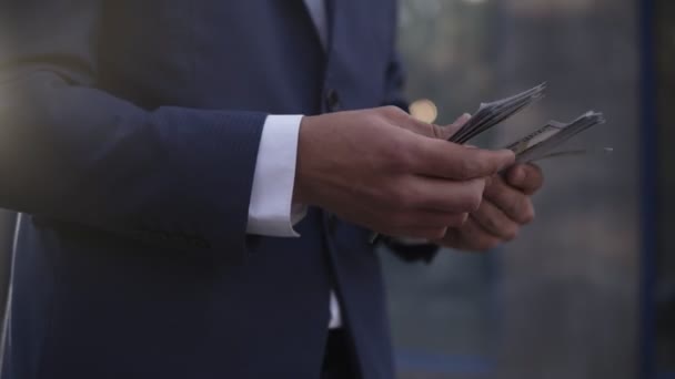 Unrecognizable businessman on the street holding bundle of american money in his hands, counting it. Wealthy male counts hundred banknotes, wearing blue suit. Close up. Side view, blurred background — Stock Video