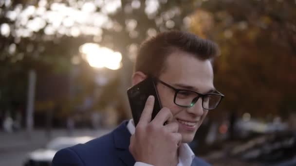 Porträtt av affärsman i snygga glasögon talar på mobilen på gatorna i affärsdistriktet. Unga leende man använder smart telefon på gatan. Solljus och höstträd på bakgrunden — Stockvideo