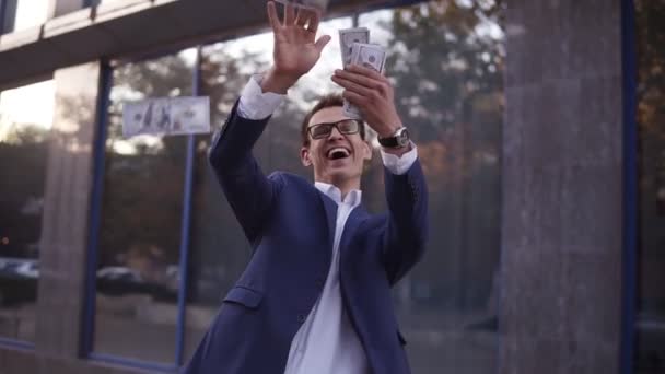 青いスーツの白人実業家は、空気中に一握りの銀行券を投げます。オフィスビルの前で現金を投げ、成功したプロジェクトと大成功を祝う眼鏡の超興奮ビジネスマン — ストック動画