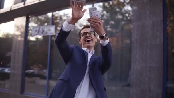 Un hombre de negocios caucásico con traje azul lanza un puñado de billetes al aire. Empresarios súper emocionados en gafas lanzan dinero en efectivo en frente del edificio de oficinas y celebran el proyecto exitoso y mucho — Vídeos de Stock