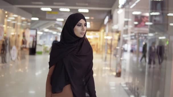 Hermosa mujer musulmana en hijab caminando por un gran centro comercial y buscando ventas a través de ventanas. Atractiva chica islámica haciendo escaparates. Movimiento lento — Vídeo de stock